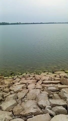 The video shows a body of water with a rocky shore. The water is calm and appears to be a lake or reservoir. The shore is covered in large, irregular rocks, some of which are partially submerged in the water. The sky is overcast, and the overall lighting is dim. There are some trees and vegetation visible on the distant shore. The camera appears to be handheld and is moving slowly from left to right, panning across the scene. There are no people or animals visible in the video. The video appears to be a landscape or nature recording.