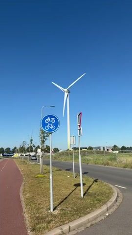 A tall white wind turbine with three blades stands prominently against a clear blue sky. In the foreground, a red paved bike path curves to the left, bordered by green grass and a grey asphalt road on the right. A blue circular sign with a white bicycle symbol is mounted on a pole next to the bike path. Further down the road, another pole holds a rectangular sign with red and white diagonal stripes. A car is visible in the distance on the left side of the image, and industrial buildings and fences are present in the background. The overall environment appears to be an open, possibly rural or industrial area, on a sunny day. There are no people visible in the frame.