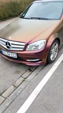 The video shows a Mercedes-Benz C-Class car parked on a street next to a curb. The car has a color-shifting wrap, appearing bronze/gold on the hood and maroon/purple on the front bumper. The license plate reads "KN-SW 111." The camera moves from the car to the pavement of the street. The lower portion of the person filming is visible. They are wearing blue jeans and white sneakers. The environment includes a street with painted lines and greenery in the background.