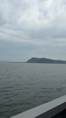 The video shows a wide expanse of choppy gray water under a cloudy sky. In the distance, a large, flat-topped hill or mountain rises from the horizon, its slopes covered in green vegetation. The foreground is partially occupied by a gray railing, suggesting the viewpoint is from a boat or similar vessel. The camera is stationary, and there is no discernible movement other than the gentle rocking of the water. The overall atmosphere is calm and somewhat muted, consistent with overcast weather.