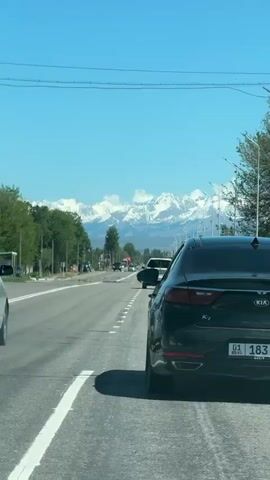 The video shows a view from inside a car moving along a highway. Several other cars are visible, along with trees, streetlights, and power lines along the side of the road. In the distance, there's a mountain range with snow-capped peaks under a clear blue sky.  The car in front of the viewer is a black Kia K7, identified by the emblem on the rear.
