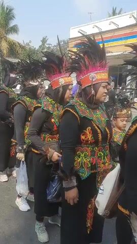 A group of people in traditional attire are standing in a line. They are wearing black clothing with colorful embroidered vests and feathered headdresses. Some are holding plastic bags. The background shows a street with trees and buildings, including an Indomaret sign. The sky is clear and the sun is shining.