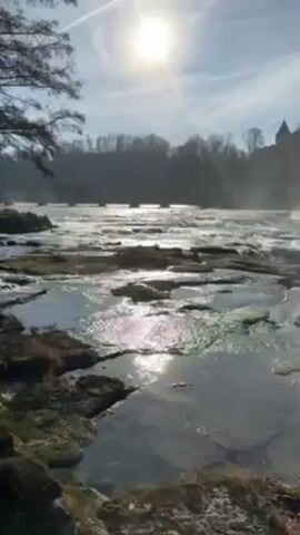 The video shows a rocky riverbed with a bright sun overhead. Water cascades over the rocks, creating mist that partially obscures a large stone building on a hill in the background. The camera pans to the right to better reveal the building. The sun reflects brightly off the water's surface.