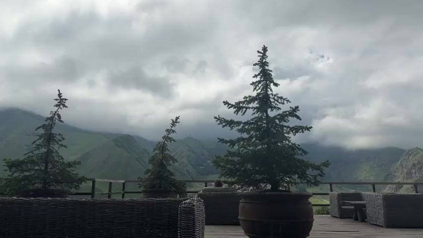 The video opens on a cloudy day with a view of a mountain landscape and some trees in the foreground. The camera pans to focus on a stone church with two domes situated atop a grassy hill. The church appears ancient and is set against a backdrop of misty mountains. The overall impression is one of a remote and serene location.