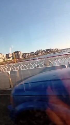 The video shows a moving perspective from inside a vehicle, likely a bus or car. The view is through a side window, looking out at a bridge with a decorative white railing and concrete barriers. Beyond the bridge is a body of water, and on the opposite shore are residential buildings under a clear blue sky. Cars pass by on the bridge. A road sign indicating a weight limit of 8 tons is visible.