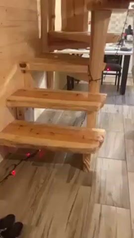 The video shows the interior of a cozy cabin. There are custom-built wooden stairs with red Christmas lights. A hammock hangs from the exposed ceiling. A brick pillar houses a wood-burning stove with a fire visible inside. A TV mounted on the pillar shows a scene from a movie.  A gray sofa sits against a wood-paneled wall, along with a chair and various board games. The kitchen area is visible in the background.