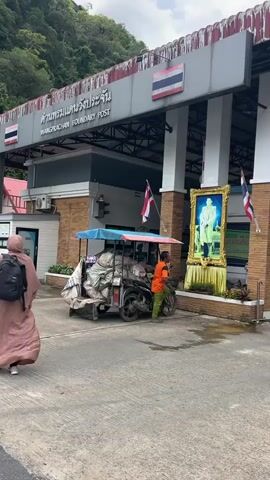 The image shows the Wangprachan Boundary Post, a border crossing in Tambon Wang Prachan, Thailand. The sky is overcast, suggesting a cloudy day. A man in an orange shirt and green pants is walking towards a motorcycle with a sidecar loaded with large, covered bundles. The motorcycle is parked in front of a building adorned with a large framed portrait of a man in a military uniform, likely a former King of Thailand. Two Thai flags are visible, one on the building and one mounted on the motorcycle's sidecar. The building's facade is a mix of brick and concrete, with a prominent sign in Thai and English reading "WANGPRACHAN BOUNDARY POST". To the left, a person wearing a hijab and a pink backpack walks away from the camera. The scene appears to be active, with people and vehicles present at the border crossing. The overall impression is one of a functional border checkpoint, with elements of national pride and daily life.