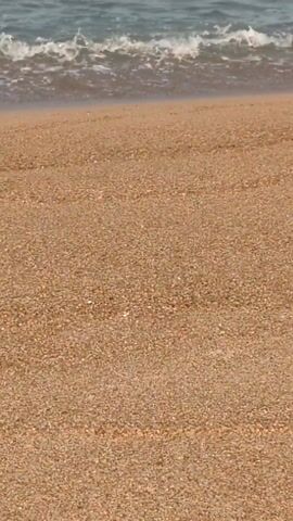 The video shows a beach scene. The focus is on the sand, which is a light tan color and appears to be made of small pebbles. In the background, the ocean is visible with small waves coming towards the shore. A crab is partially buried in the sand, near a small hole.