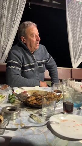 A man sits at a table laden with food and drinks. He is wearing a zipped-up gray sweater with white stripes on the chest and arms. The table is covered with a blue and white patterned tablecloth. In front of him is a plate with food, ashtray, lighter, and glasses. Behind him, a white curtain is visible. The background is dark, indicating it is nighttime.