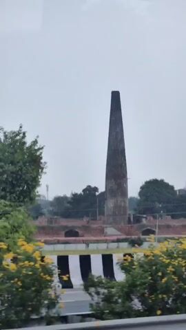 A tall, narrow, conical brick chimney stands against an overcast sky. The chimney appears weathered and aged. It is surrounded by greenery, including trees and bushes. In the foreground, a black and white striped barrier separates the viewer from a road. Beyond the chimney, other buildings and structures are visible, some partially obscured by trees. The overall impression is of an industrial or historical site within a vegetated landscape.