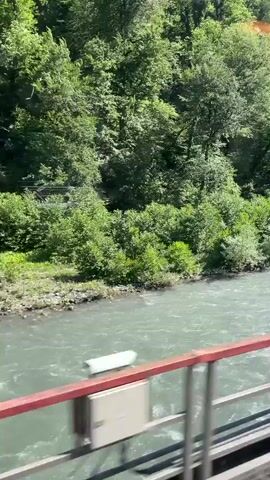 A river flows swiftly through a wooded valley. The camera is looking out from what appears to be a bridge or elevated walkway, as a railing with red and metal accents is visible in the foreground. The river's water is a cloudy, pale green and shows signs of movement with ripples and small waves. The banks of the river are covered in lush green foliage, with trees densely packed on the sloping hillsides that rise sharply behind the river. Sunlight filters through the leaves, creating dappled patterns and a bright, sunny atmosphere. There are no people or animals visible in the frame.