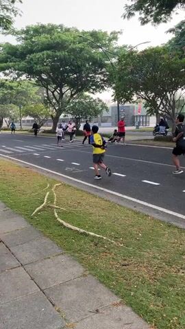 A group of people are engaged in various outdoor activities on a paved path beside a road lined with trees. In the foreground, a person on a skateboard moves from left to right. To their right, a person runs on the road. Further down the road, several other individuals are running, walking, or standing. One person is seated on the ground. On the far right side of the road, another person walks. A cyclist on a blue bicycle is seen on the road among the pedestrians. The environment appears to be a park or a well-maintained urban area, with green grass bordering the path and large trees providing shade. A building is visible in the background. The lighting suggests it is daytime, possibly early morning or late afternoon.
