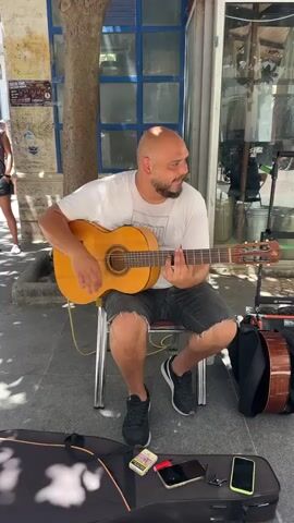 Two men are playing acoustic guitars outside a store. The man on the left is bald, wearing a white t-shirt and dark shorts. He is sitting on a metal chair and playing a light brown acoustic guitar. A black guitar case is open on the ground in front of him, with a pack of cigarettes, two cell phones, and a pair of sunglasses on it. There is also a small black amplifier with orange trim behind him. The man on the right is wearing a dark t-shirt, shorts, and sunglasses. He is sitting on a black metal chair and playing a red acoustic guitar. A black guitar case is behind him. There is a water bottle on the ground in front of him. The ground is paved with cobblestones. The store behind them has large glass windows. Other people are walking by in the background. The scene appears to be on a sunny day.