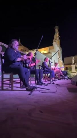 A dimly lit outdoor performance area at night is illuminated by purple stage lighting. Four men are seated on chairs on a wooden stage. The man on the far left is clapping his hands. The second man from the left is also clapping his hands and appears to be singing into a microphone. The third man from the left is seated, wearing a suit jacket, and has his hands clasped in his lap. He is also facing a microphone. The man on the far right is seated and playing a guitar. A building with a tall spire is visible in the background, illuminated by warm light. There are microphones and cables on the stage.