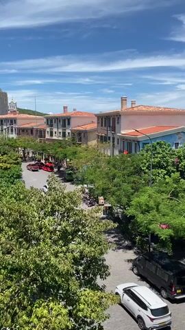 This media file presents a serene, daytime scene in Phú Quốc, Vietnam. The setting is a charming street lined with buildings featuring terracotta roofs and Mediterranean-inspired architecture, suggesting a resort or upscale residential area. Lush green trees with vibrant foliage are abundant, casting dappled shadows on the cobblestone street below, indicating a pleasant and warm climate.

The street is moderately active with various vehicles. A black van and a white SUV are parked, while a distinctive bright red open-top vehicle, possibly a tourist tram or a novelty car, is driving along the road. In the distance, a few other vehicles, including a red one, can be seen, along with what appears to be a few people near some buildings. The overall atmosphere is peaceful and relaxed, with the bright blue sky dotted with fluffy white clouds suggesting a beautiful, sunny day, likely mid-morning or early afternoon. The distant hint of the ocean implies the proximity to the coast, a common feature of Phú Quốc. No text is visible in the media file.