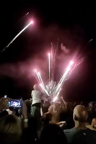 This is a video of a fireworks display at night. A crowd of people are gathered to watch. In the foreground, several people are holding up their phones to record the event. A child is being carried on someone's shoulders to get a better view. The fireworks are exploding in various colors, including pink, green, and white, creating a dramatic and festive atmosphere. The sky is dark, and smoke from the fireworks is visible. Some of the fireworks are creating trails of light as they ascend and descend. The overall scene is one of excitement and celebration.