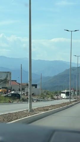 The video, taken from inside a moving vehicle, showcases a picturesque scene of a highway with vehicles driving on it, set against a backdrop of mountains under a bright blue sky with scattered clouds. The road appears to be under construction, with some unfinished portions and construction equipment visible on the side. There are several lampposts lining the road. In the distance, the mountains are covered with lush greenery. The vehicles include a bus, a white van, and other cars. There are also signs and billboards alongside the road. The overall impression is that of a highway journey through a scenic mountainous area.