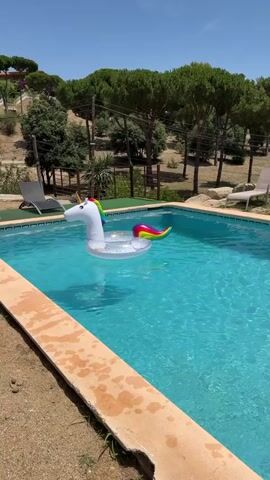 A white unicorn-shaped inflatable float is shown in a swimming pool. The pool has blue water and is surrounded by a concrete deck and sandy area. In the background, there are green trees, a blue sky, and some lounge chairs. The video is shot from a slightly elevated angle, looking down at the pool. The float is the main subject of the video and is positioned in the center of the pool.