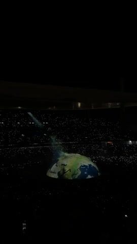 The scene is very dark and grainy, set inside a large stadium. A domelike structure displaying an image of the Earth is positioned in the center, illuminated by a spotlight. There is a person standing atop this Earth projection. The background is filled with a sea of small white lights, likely from mobile phones held up by people in the audience. The stadium's roof structure is dimly visible at the top of the frame.