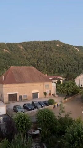 The video shows a view from a high vantage point overlooking a rural village. In the foreground, there is lush green foliage and what appears to be a patio or deck area. Dominating the midground is a large, light-colored building with a reddish-brown roof. In front of this building, there is a parking area with several cars, including at least four dark-colored SUVs and one lighter-colored SUV. A street curves away from the building and disappears into the distance. The background is dominated by a steep, densely wooded hillside. The sky is a clear, pale blue, suggesting it is either early morning or late evening. There is a small, slender structure, possibly a communication tower, visible on the crest of the hill in the distance. The overall impression is one of a peaceful, somewhat remote setting.
