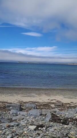 This media file captures a serene coastal scene along Pier Road in Ireland. The foreground is dominated by a rocky shoreline strewn with various pebbles and larger grey stones, interspersed with dried seaweed. Beyond the rocks lies a strip of light-colored sand, the transition zone between land and sea.

The main subject is the vast expanse of the ocean, its surface a deep blue with gentle ripples reflecting the sky. The water appears calm, suggesting a tranquil day. In the distance, a long, flat island or peninsula stretches across the horizon, its form softened by a hazy layer of clouds that hug the waterline. Further inland, the land rises gently.

The sky above is a dynamic canvas of bright blue interspersed with dramatic white clouds. Some clouds are wispy and streaked, while others are more voluminous and textured, adding depth to the scene. The interplay of sunlight and cloud cover suggests it is daytime, likely late morning or afternoon, with fair weather. The overall atmosphere is peaceful and natural, evoking a sense of quiet contemplation and the beauty of the Irish coast. There is no visible text in the media file.