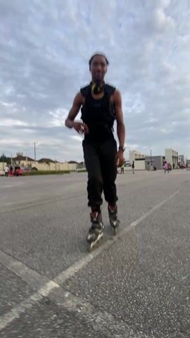 Here's a description of the video:
The video shows a man inline skating on a gray asphalt surface. He is wearing black pants, a black sleeveless shirt, and a black vest. He has dark hair in dreadlocks and is wearing a watch on his left wrist. He is moving towards the camera. The sky is overcast, and there are buildings and cars visible in the background. Other people appear to be roller skating as well. The camera angle is low, focusing on the man's legs and skates as he moves.