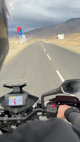 The video shows a first-person perspective of someone riding a motorcycle on a paved road. The rider's hands are visible on the handlebars, and the motorcycle's digital display shows the speed decreasing from 99 to 88. The road is flanked by dry, barren land and mountains in the background. The sky is cloudy with dark storm clouds. There are a few people and vehicles visible in the distance. The overall impression is a solitary journey through a remote, mountainous landscape.