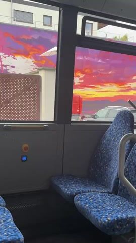 This video shows the interior of a bus. The camera is positioned to capture a view through the bus windows. The seats inside the bus are blue with a speckled pattern. Outside the window, a vibrant mural depicting a sunset with intense shades of orange, pink, and purple is visible on the side of a building. In front of the mural, a red lift truck is parked, and a car is visible in the background. The overall environment appears to be a city street.