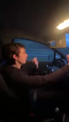 The video shows a young man driving a car at night. He is wearing a dark sweatshirt and pants. He is moving his hands as if drumming in time to the music. The interior of the car is visible, including the dashboard, steering wheel, and passenger seat. Through the window, you can see the road and some trees in the distance. The lighting is dim inside the car, with only the overhead light providing illumination.
