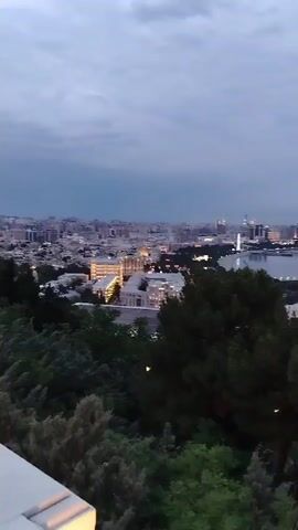 The video shows a panoramic view of a city skyline at dusk, overlooking a body of water and a densely forested area in the foreground. The sky is overcast with a pale blue and grey hue. The city lights are beginning to illuminate, with several tall buildings and a distinctive curved structure visible in the distance. The water appears calm. The foreground consists of lush green trees and shrubs, with some appearing to be coniferous. The lighting suggests it is either early morning or late evening. The camera angle is elevated, providing a wide-angle perspective of the cityscape.
