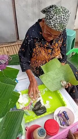 An elderly woman wearing glasses and a headscarf is seated, preparing food. She is dressed in a dark patterned blouse and a dark skirt. Her hands are actively engaged in wrapping something in banana leaves. In front of her, on a table, are several items including green banana leaves, a green tray with white shredded material and some dark food items, a small white bowl with colorful sprinkles, a pink basket filled with triangular-shaped food items wrapped in leaves, a red container, a small bottle with a brown liquid, and some tools. The setting appears to be outdoors or in an open-air market, with a corrugated metal wall in the background and a green plastic stool to the right.