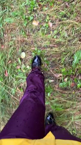 The video shows a first-person perspective of someone walking through tall green and brown grass, with some scattered leaves and dirt visible on the ground. The person is wearing dark purple rain pants and black boots. The ground appears wet, suggesting it might be raining or has recently rained. The camera is moving forward with each step, indicating the person is actively walking. The overall environment appears to be outdoors, possibly a park, trail, or unkempt yard.