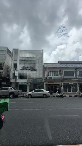 The media file captures a street scene in Phuket City Municipality, Thailand, under a cloudy sky. The view is from the perspective of someone on the street, looking towards a row of buildings lining the road.

The primary subjects are the buildings themselves, which showcase a mix of architectural styles. On the left, a white building with a distinctive perforated facade stands out. It features a sign that reads "BumBumRoom" in a stylized font, along with other smaller text. Adjacent to it, to the right, is a building with a more traditional facade, featuring windows and signage. Further to the right, another building with a similar style is visible, including a sign with Thai script and an image of a person.

Several objects are present on the street. Cars are parked along the road and driving by, including a silver sedan and a white pickup truck. A motorcycle with a green delivery box is partially visible in the foreground. Scooters are also parked in front of some of the shops.

The scene appears to be an urban commercial area, with storefronts suggesting various businesses. The cloudy weather creates an overcast atmosphere, and the time of day is not precisely discernible but appears to be daytime. No specific activities or events are clearly depicted beyond the general movement of vehicles and the presence of businesses. The overall mood is neutral, reflecting a typical street view.

Visible text includes "BumBumRoom," Thai script, and other smaller signs on the storefronts, likely indicating the names or services of the businesses.
