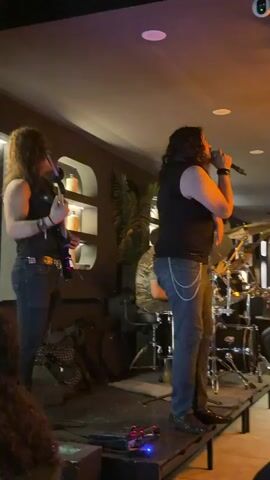The video shows a live band performing on a stage. Two male singers are prominently featured. The singer on the right has long, dark hair, is wearing a black tank top and jeans, and is holding a microphone. He is gesturing with his free hand and appears to be singing energetically. The singer on the left also has long, dark hair and is wearing a black tank top and jeans. He is holding a guitar and appears to be singing into a microphone. A drummer is visible in the background, seated behind a drum kit. The stage is covered with a dark material, and there are speakers and other electronic equipment on the floor in front of the stage. The background of the venue is dimly lit, with some shelves and decorative items visible on the wall. The overall atmosphere suggests a rock or metal music performance.