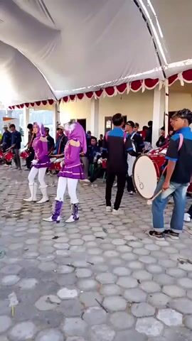The video shows a group of people practicing for a marching band. Three individuals in purple and white uniforms with headscarves are marching in the foreground, holding batons. Several other individuals are behind them, some holding drums and other instruments. They are practicing under a white tent with red trim. The ground is paved with hexagonal stones. The setting appears to be outdoors.
