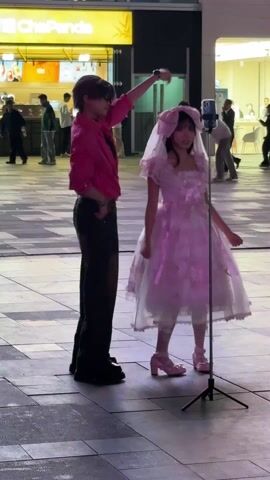 The scene shows two people, one a young girl wearing a pink frilly dress and a headdress, and the other a young man in a pink shirt and dark pants. They are standing in a public square. The girl is wearing pink shoes and has a cat tail attached to the back of her dress. The man has his arm around her, apparently adjusting the headdress or hair. They are standing in front of a smartphone on a tripod, presumably recording a video. In the background, there are several pedestrians and a restaurant named ChePanda. The setting appears to be a well-lit, outdoor urban area.