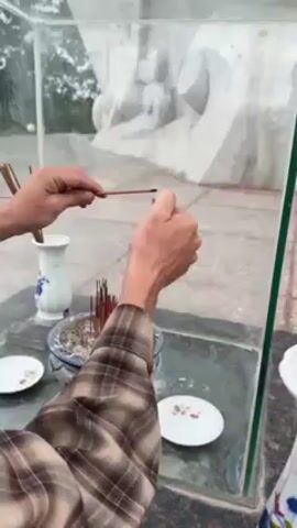 A person wearing a plaid shirt uses a lighter to light an incense stick. They hold the incense stick in their left hand and the lighter in their right. The incense stick ignites and a small flame appears at the tip. In the foreground is a glass-covered memorial or altar area. Within the enclosed space are several small white plates, a vase, and a pot filled with sand and more incense sticks. A large white statue can be seen through the glass enclosure. The surrounding environment is outdoors.