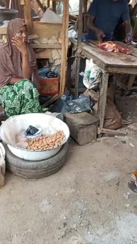The video shows a street market scene. A man in a blue shirt is cutting meat at a wooden table.  A woman in a green and orange patterned skirt is walking away from the table, carrying an orange bucket.  Another woman in a red dress walks by, carrying a blue plastic bag and an orange bucket. Other people pass by various stalls with goods displayed.  The environment is dusty, and the market appears to be busy.