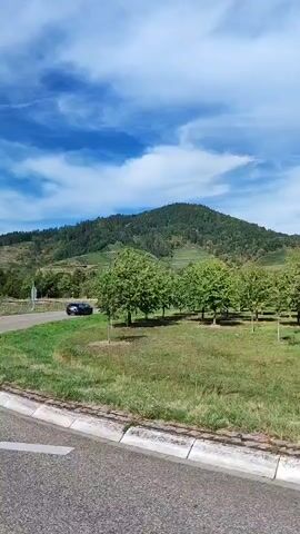 A car is seen driving on a road with green trees and hills in the background under a blue sky with white clouds. There are signs indicating directions to Oberkirch-Ost and U2. The video appears to be shot from a moving vehicle.