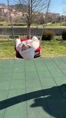 A toddler, bundled in a white, hooded jacket and maroon pants, swings in a red-framed, black-seated swing at a playground. The swing is suspended by chains, and the playground has a green tiled surface. In the background, there is a grassy area, bare trees, a stream, benches, and buildings in the distance under a clear blue sky. The toddler appears to be enjoying the swing.