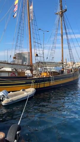 This media file depicts a beautiful, large wooden sailing ship docked in a harbor under a clear blue sky. The ship is painted black with a distinctive yellow stripe along its hull. It has two tall masts with a complex rigging system, indicating it's a traditional schooner.

Tied to the side of the main ship is a smaller, inflatable dinghy with a Honda outboard motor. The dinghy is white and appears to be used as a tender for the larger vessel. The water is calm and reflects the blue sky and the ship.

In the background, more boats and buildings can be seen along the waterfront, suggesting a busy port town. The architecture of the buildings hints at a Mediterranean setting, which is further confirmed by the provided location context of Palamós, Spain. A modern, multi-story building stands out in the distance, contrasting with the historic nature of the sailboat.

Several flags are flying from the ship's rigging, though the text on them is largely illegible, except for one banner that appears to have "WAY" written on it. There are also a couple of life preservers visible on the deck of the ship.

The overall impression is one of a bright, sunny day, likely in the late morning or early afternoon, given the strong sunlight and clear conditions. The scene evokes a sense of maritime heritage and leisure, suggesting the ship is either a historical vessel, a charter boat, or a private yacht. No specific activity or event is clearly taking place; the ship appears to be at rest in the harbor.