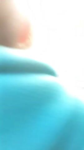 The video shows a first-person perspective of someone riding a scooter on a paved road. The sun is low in the sky, casting a warm glow and creating lens flare. On the right side of the scooter's handlebar, a mirror reflects the rider's green hoodie. The road appears to be in a rural or suburban area, with houses and trees visible on either side. Other scooters and people can be seen further down the road. The scooter has a black and silver dashboard with a speedometer. The rider's hands are visible on the handlebars. The overall mood is peaceful and serene, with the golden hour light enhancing the natural beauty of the scene.
