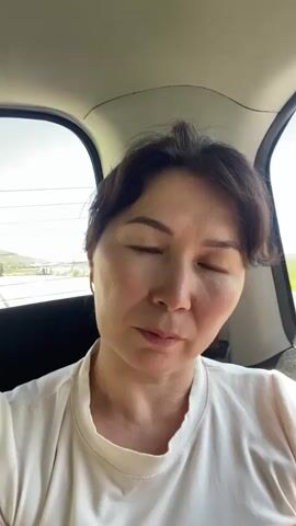 A woman with dark hair and light skin is sitting in the back of a car. She is wearing a light-colored shirt. The car appears to be in motion. The background shows a blur of green fields and a road with other vehicles. The woman blinks and smiles at the camera.