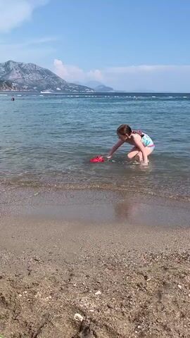A young girl in a colorful swimsuit walks out of the ocean holding a red bucket with a green handle. The water is clear and blue, with small waves lapping at the sandy shore. In the background, mountains rise against a bright blue sky with scattered clouds. The girl is looking towards the viewer.