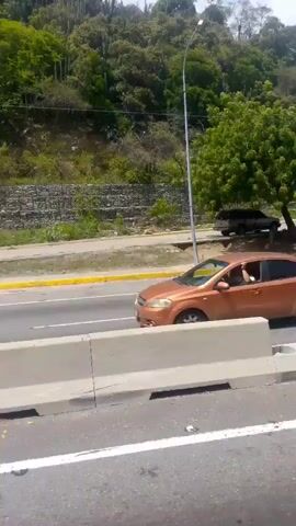 The video shows a view from a moving vehicle on a road in Macuto, Venezuela. The foreground consists of the road itself, with clear white lane markings and a concrete barrier separating the lanes. Beyond the barrier, there is a sidewalk with some debris, and further back, a hillside covered in lush green vegetation. A street light stands tall on the right side of the frame, and a large, leafy tree partially obscures a parked dark-colored car. The road appears to be a highway or a major thoroughfare, with multiple lanes visible. The scene suggests it is daytime, likely sunny, given the bright lighting and clear visibility. The overall impression is of a typical roadside scene in a tropical or subtropical environment. There is no discernible activity or event taking place beyond the movement of the vehicle from which the video is being recorded. No text is visible in the frames.