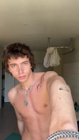 A medium shot shows a shirtless young man standing in a bathroom. He has brown, wavy hair and wears a silver chain with a pendant and a silver bracelet. He has a small tattoo on his upper chest and another on his upper left arm. He is wearing blue and white patterned swim trunks. The background shows a white claw-footed bathtub, a white shower curtain, a white cabinet, and white tiled walls. The man runs his hand through his hair, then looks at the camera and claps.