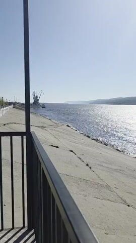 The camera pans right from behind a black metal railing along a concrete embankment to reveal a wide body of water sparkling in the sun, with a treelined shore in the distance. A crane is visible on the left.