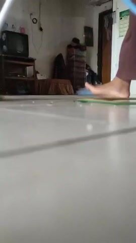 A person's bare feet and the bottom of their brown pants or skirt are visible as they sweep a light-colored tiled floor with a broom that has a green and yellow head and a silver handle. The broom pushes small specks of debris across the floor. In the background, to the left, a blue laundry basket filled with clothes is visible next to a light-colored wall. Further back and to the left, a small black television sits on a wooden stand. The camera is at a low angle, close to the floor.