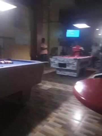 The scene depicts the interior of a pool hall. There are two pool tables visible: one with a blue felt top and balls racked at one end, and another with a red felt top and a white and grey camouflage-patterned base. On the camo table, a man wearing a red shirt is lining up a shot, while two other men stand nearby, observing. Another man is standing near a wall in the background. A red round table is also visible in the foreground. A television is mounted on the wall above the camouflage table and shows a sports broadcast. The floor is tiled with a brown and beige pattern. The lighting is dim, creating a relaxed atmosphere.
