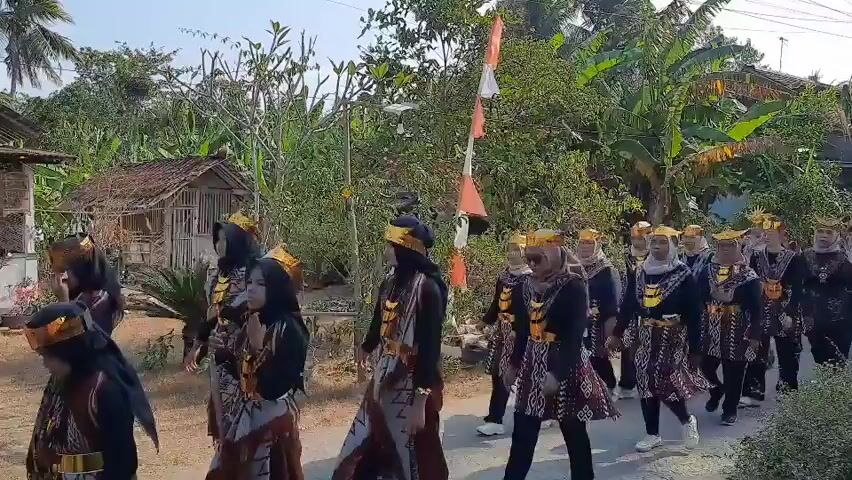 A group of people dressed in traditional clothing are walking in a procession. They are wearing black tops, patterned skirts, and gold headpieces and necklaces. The people are walking down a dirt road lined with trees and houses. There is a flag with red and white stripes flying from a pole. The sky is blue and the sun is shining. The people appear to be participating in a cultural event or celebration.